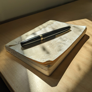 A worn pocket journal on a sunlit desk. A pocket-sized fountain pen rests atop the journal.
