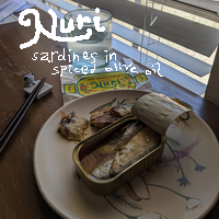 An opened can of Nuri spiced sardines in olive oil on a plate. White MS Paint lettering reads, 'Nuri sardines in spiced olive oil.'